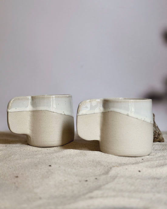 Tasse expresso Lya en grès blanc avec émaillage nacré et anse arrondie, posée sur une surface sableuse. Élégante et artisanale.
